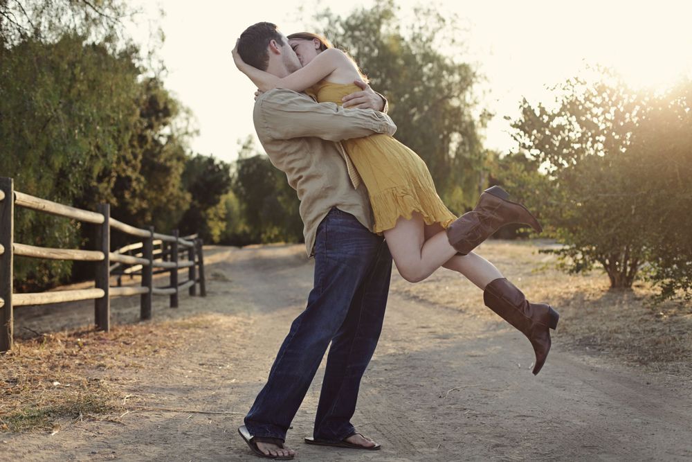 Couple embraces in playful lift pose on dirt path with wooden fence.