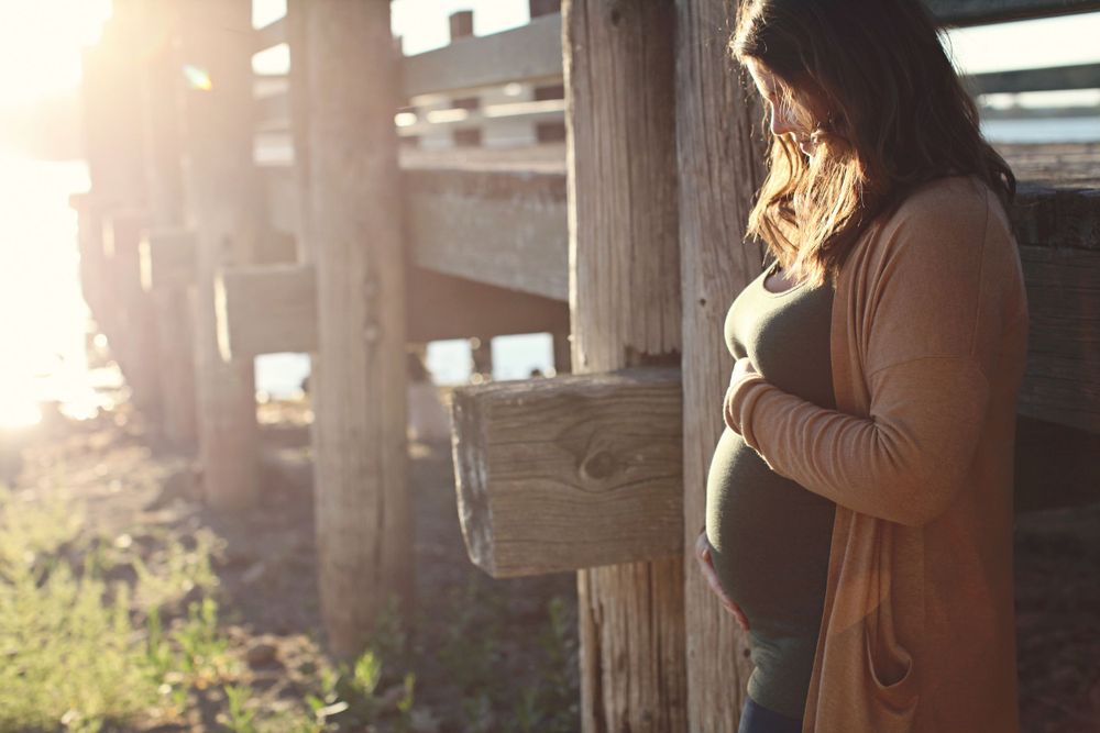 A silhouetted pregnant woman stands by wooden posts at sunset.