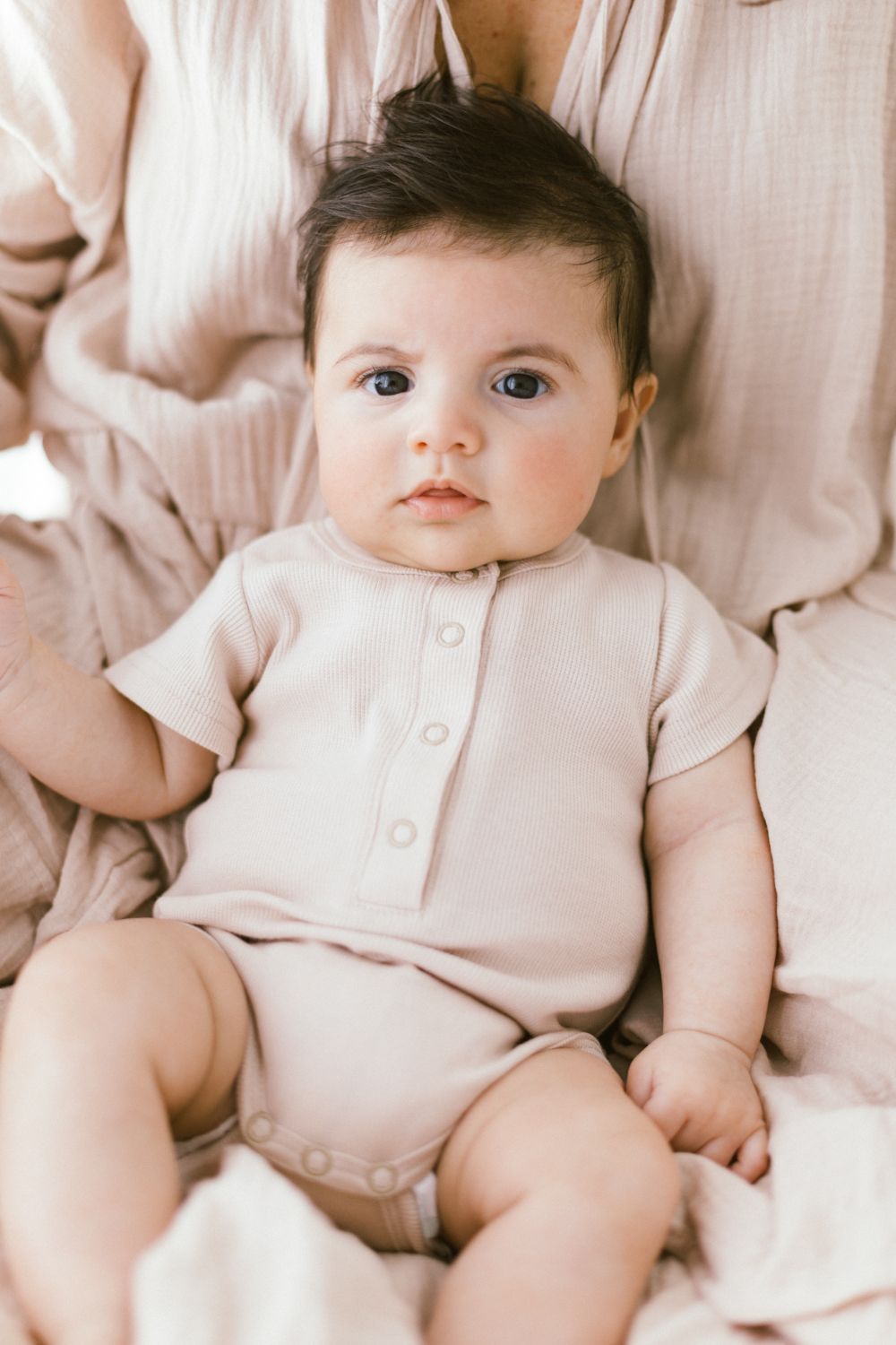 Baby in pink onesie looks directly at camera while lying on white bedding.
