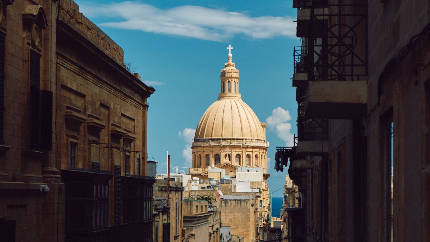 Ornate church dome with golden cross rises between historic buildings on a sunny day in Malta.
