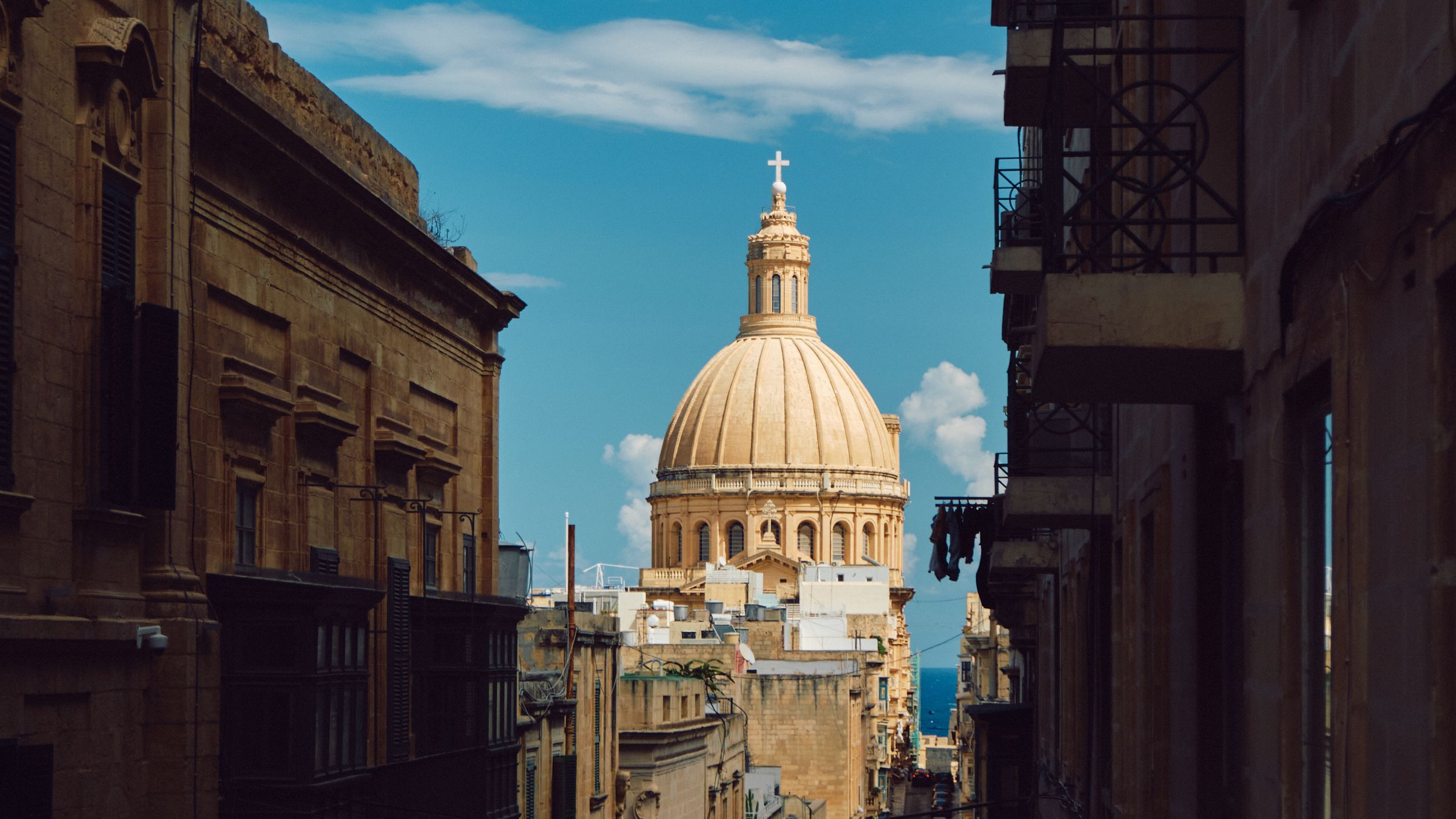 Ornate church dome with golden cross rises between historic buildings on a sunny day in Malta.