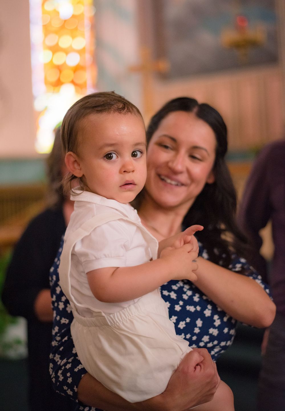 Baby in a white outfit held by an adult, looking curiously at the camera indoors
