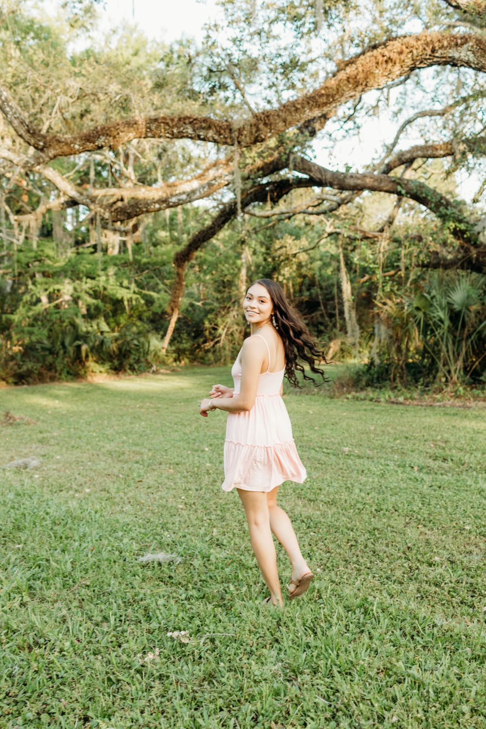 Woman in light pink dress twirls barefoot on grass under large oak tree.