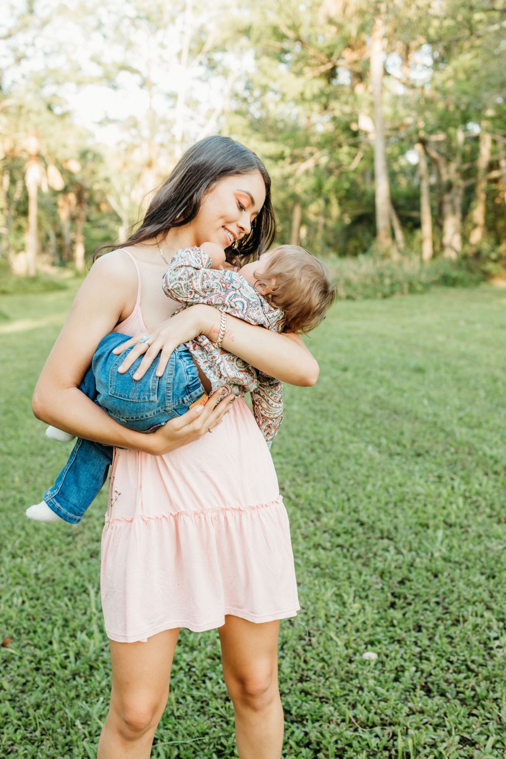 Person in pink dress holds small child wearing blue against grassy outdoor backdrop.