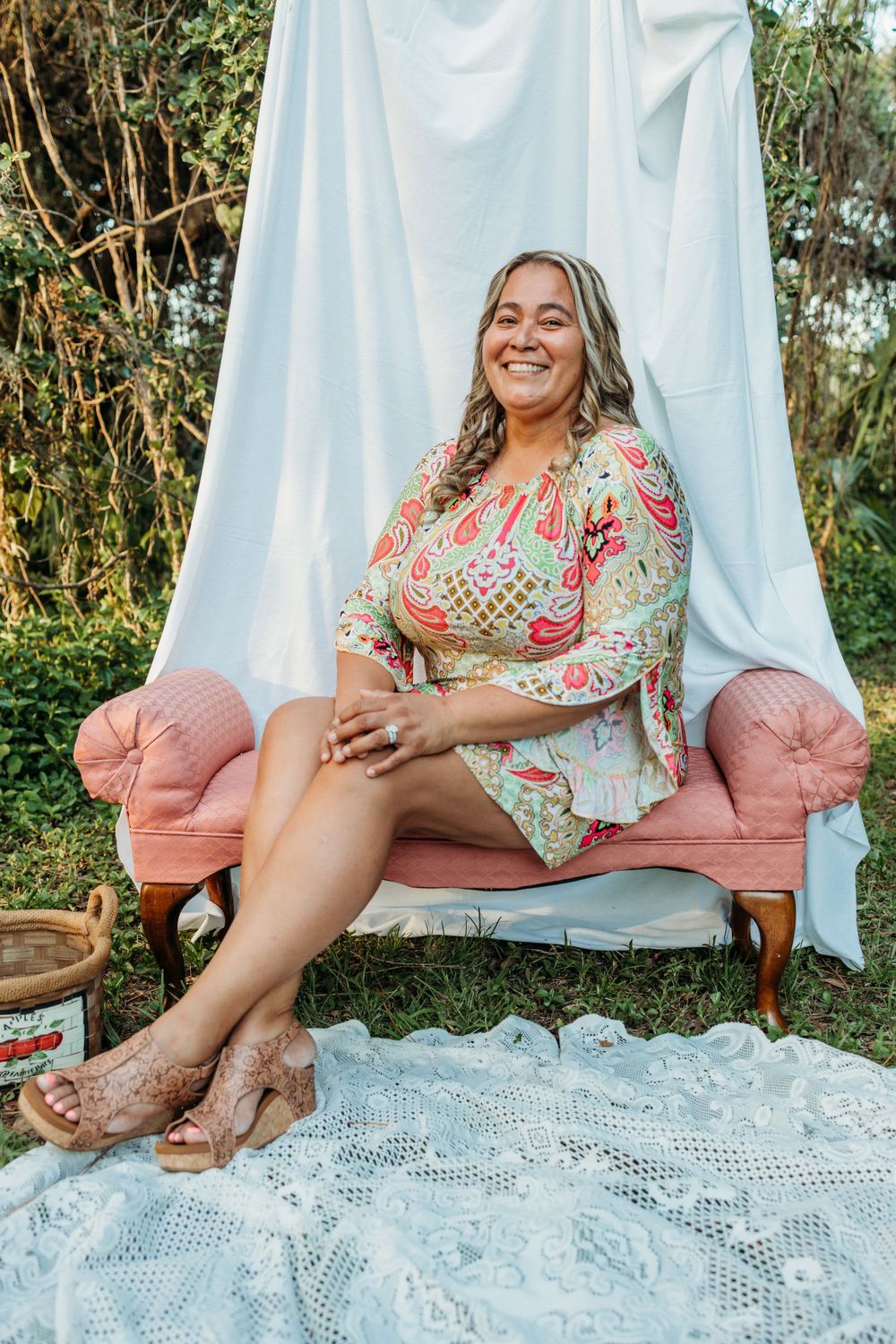 Woman in floral dress sits elegantly on pink vintage chair against white backdrop.