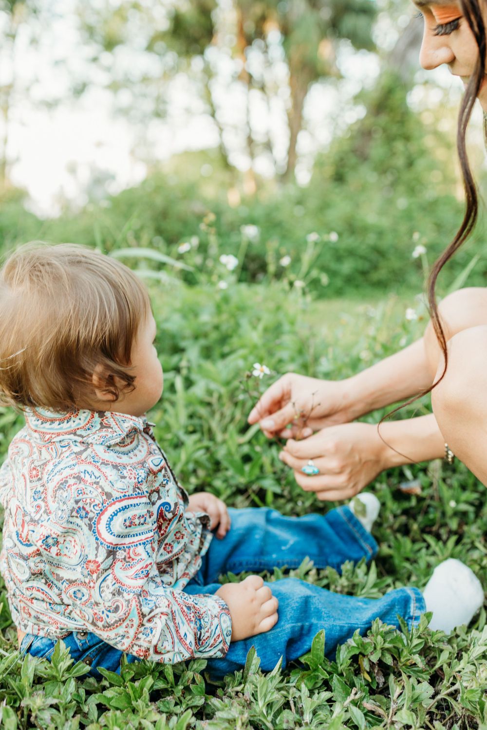 Toddler in patterned shirt sits on grass while someone offers something.