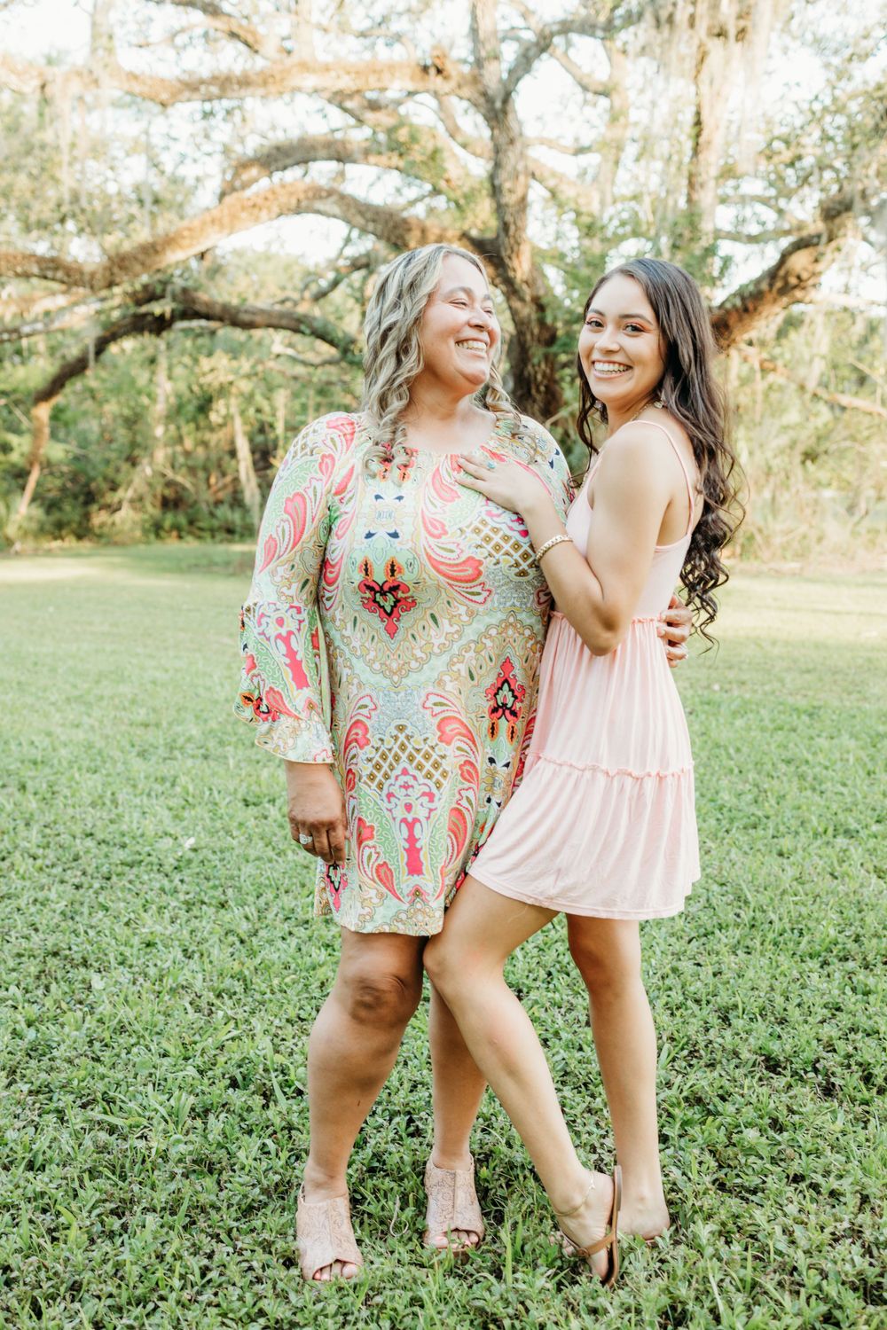 Two women in complementary dresses pose together on grass under oak trees.
