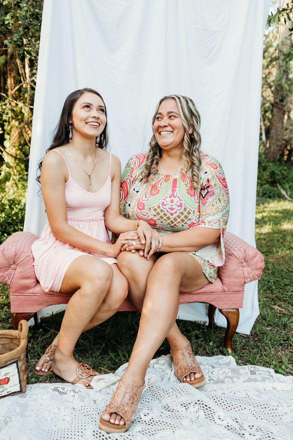 Two women share genuine laughter while sitting together on pink vintage chair outdoors.