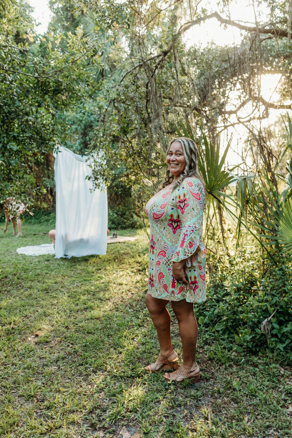Woman in floral dress poses by white backdrop in garden setting with golden sunlight.