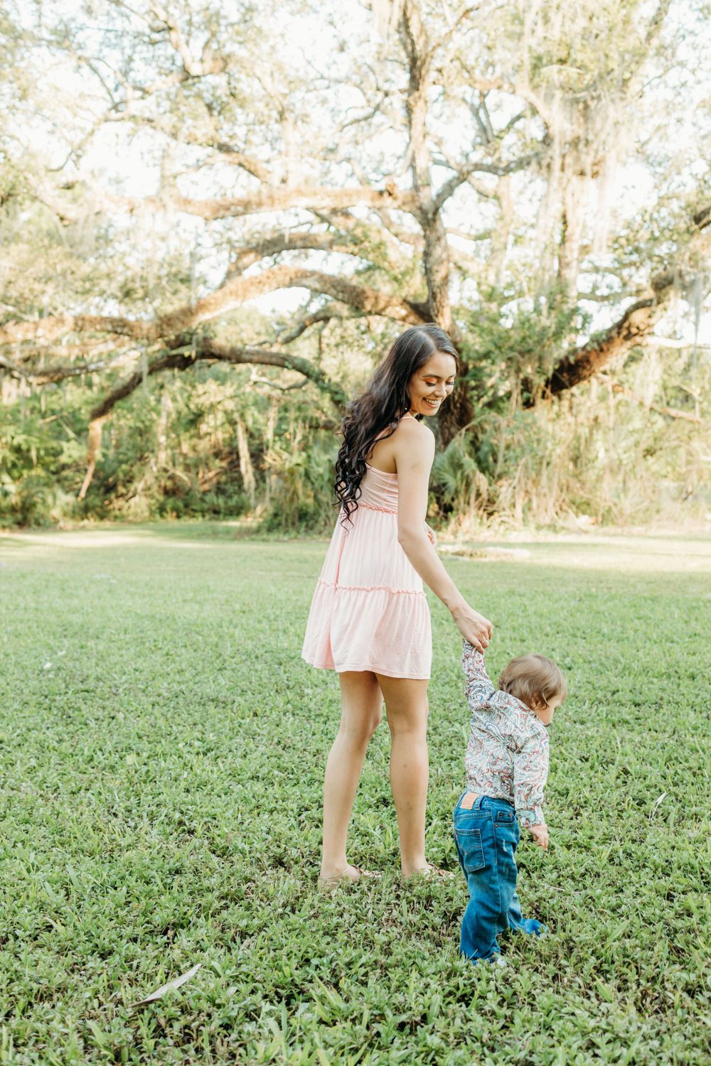 A woman in a pink dress walks with a toddler in a grassy outdoor setting.