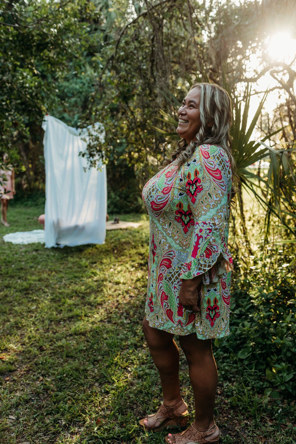 Woman in patterned dress laughs joyfully next to white fabric backdrop in garden.