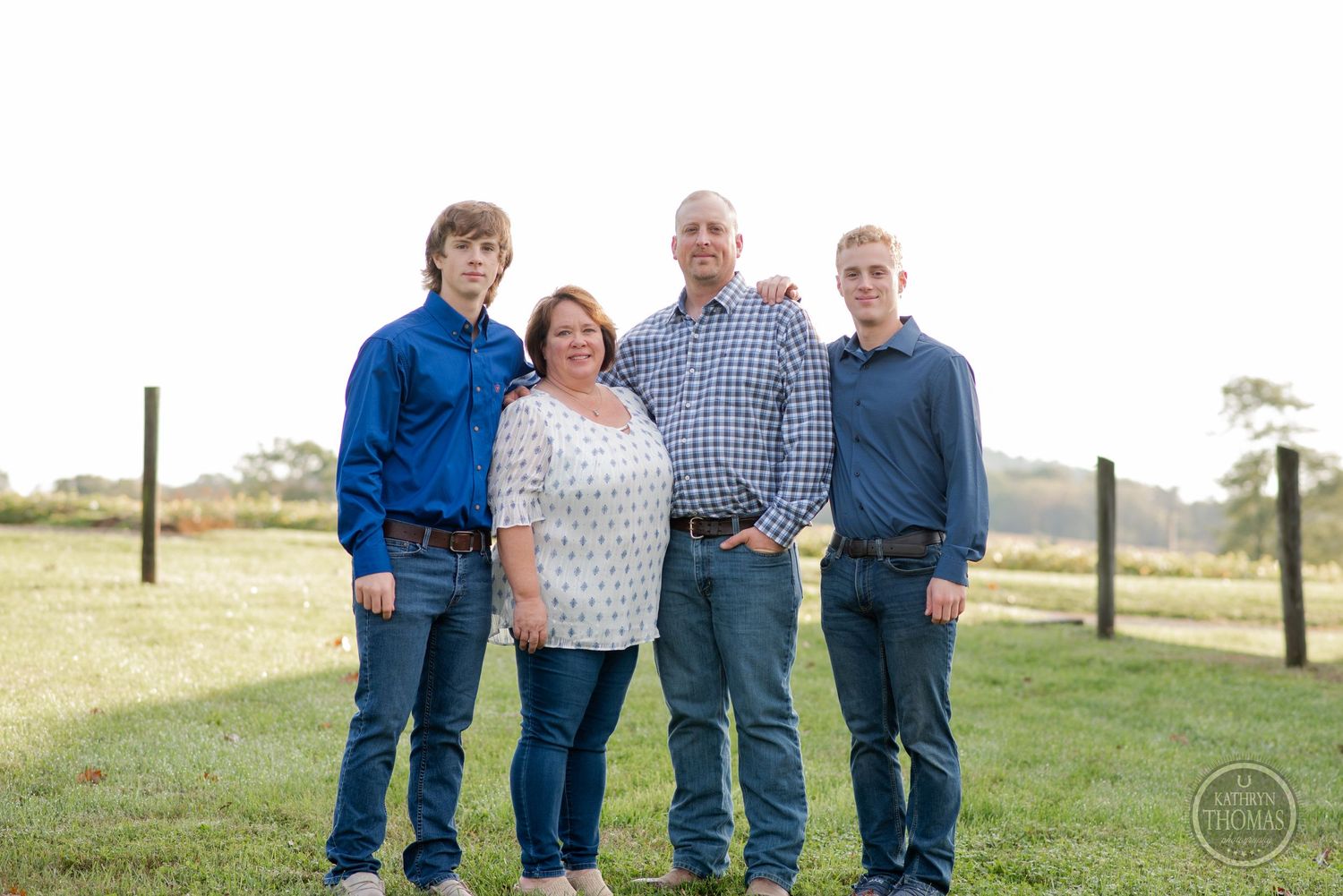 A family of four poses together outdoors in casual attire on grassy farmland.