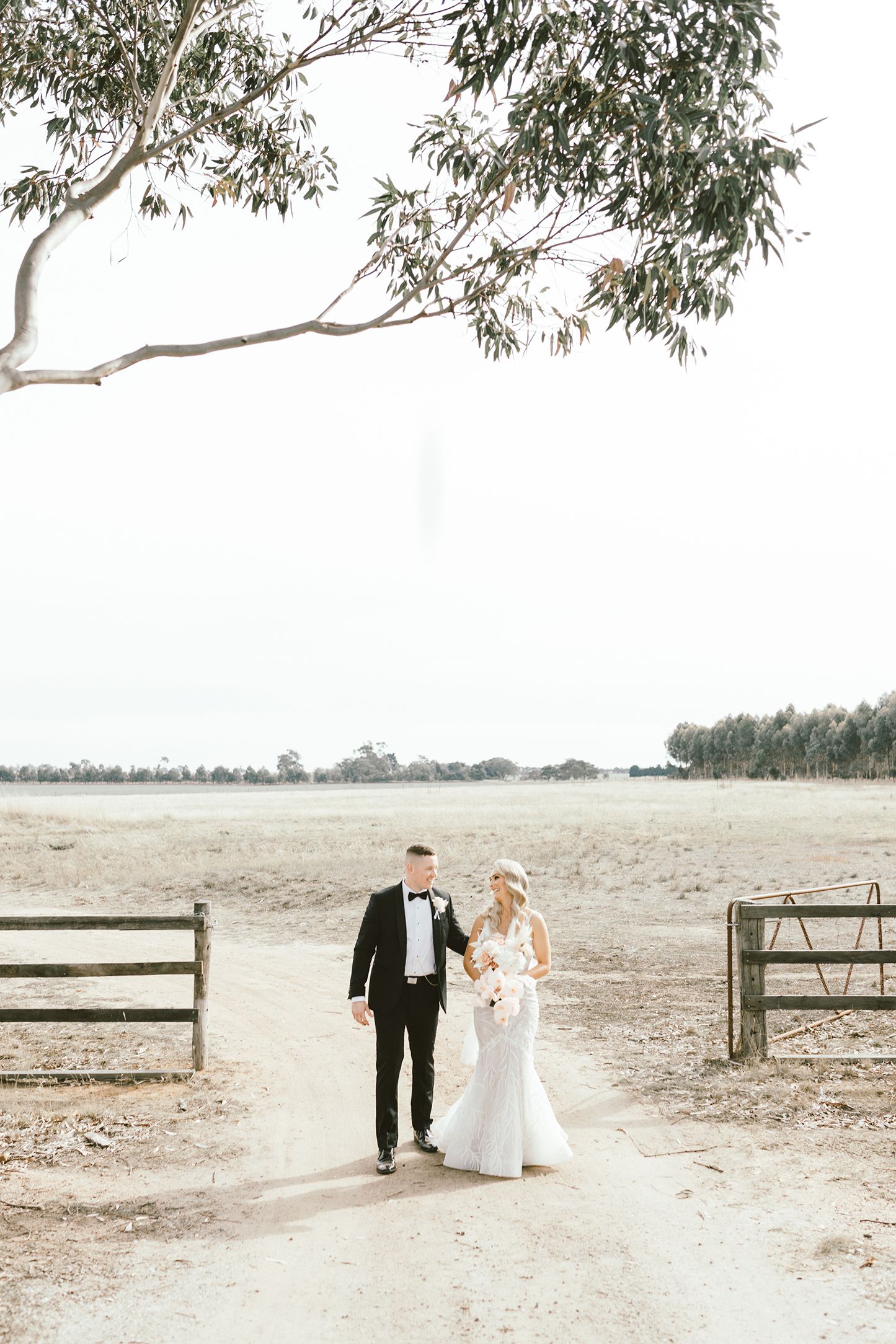 Wedding couple walking together on a countryside path with wooden fences and trees.