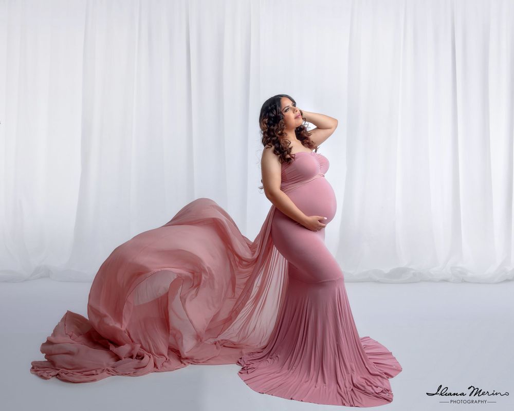 Pregnant woman in flowing pink maxi dress poses with billowing fabric against white curtain backdrop.