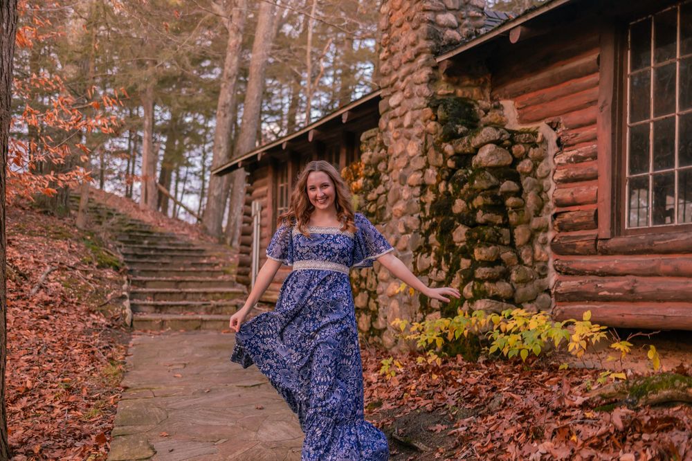 A blue floral maxi dress flows elegantly against the autumn backdrop of a rustic log cabin with stone chimney and steps.