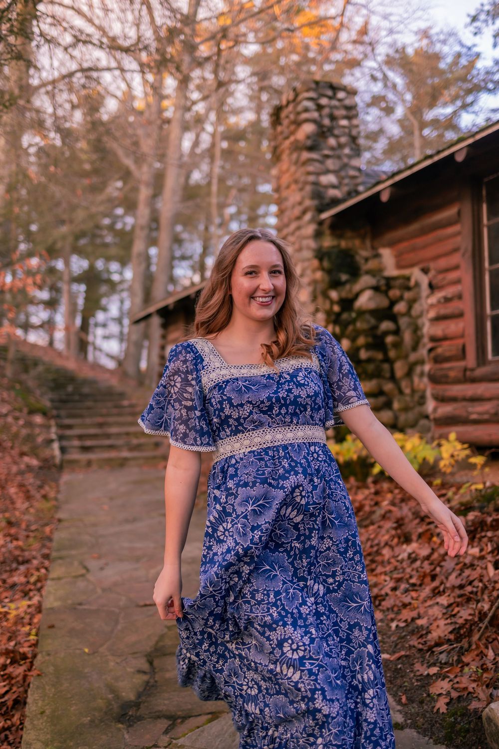 Woman wearing a blue and white floral dress poses on stone steps near rustic cabin during autumn.