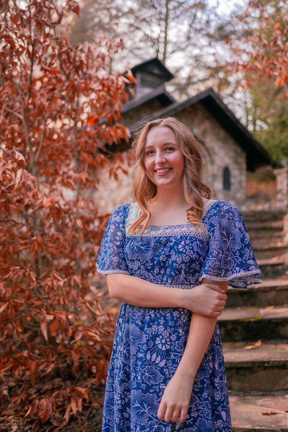 Someone in a blue floral dress poses on autumn stone steps with orange foliage and a rustic building in the background.