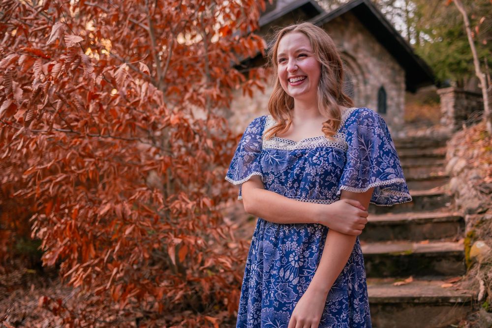 A person in a blue floral dress stands by rustic stone steps surrounded by autumn foliage.