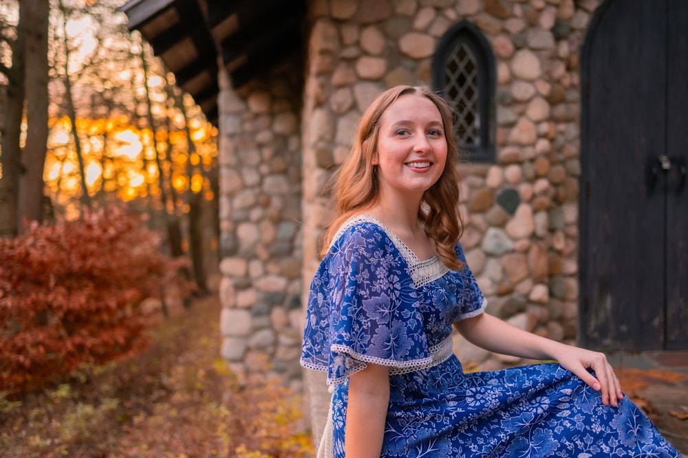 A person in a royal blue floral dress sits by a stone building during golden hour sunset with autumn foliage.