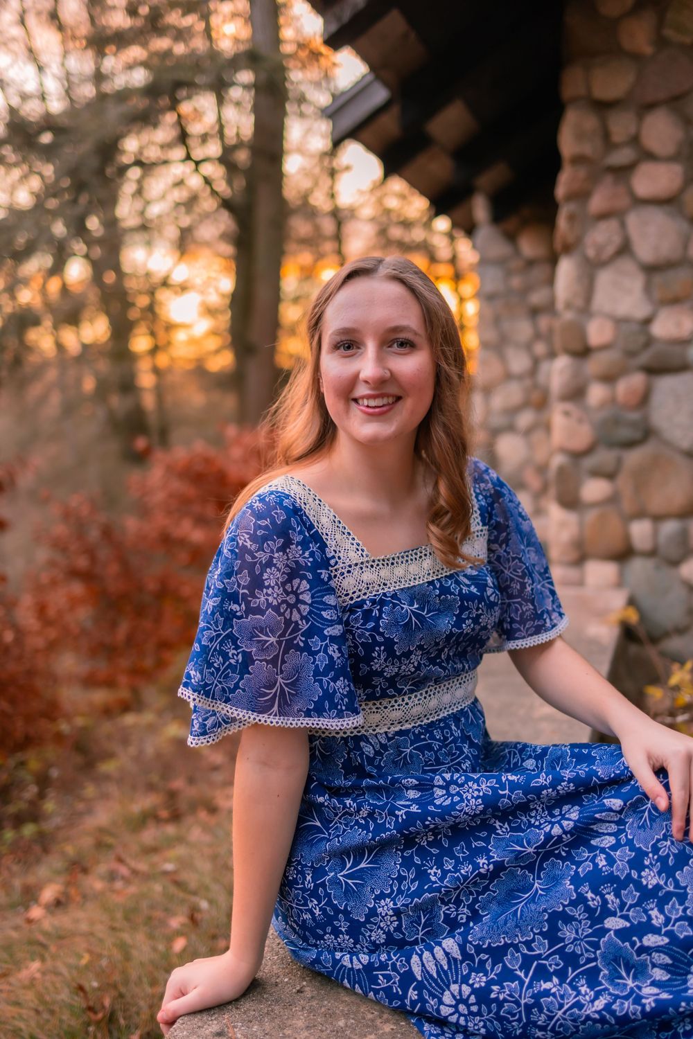Person in a royal blue lace dress surrounded by autumn foliage at sunset near a stone wall.