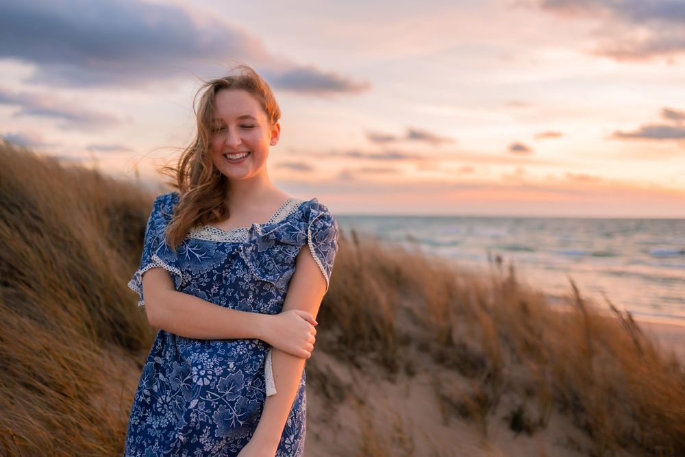 A person in a blue floral dress poses on a beach at sunset with sand dunes and ocean waves in the background.