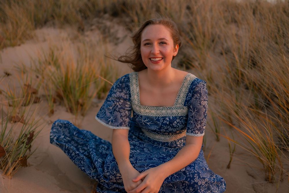 Woman in blue floral dress sits and poses on a sandy beach during golden hour.