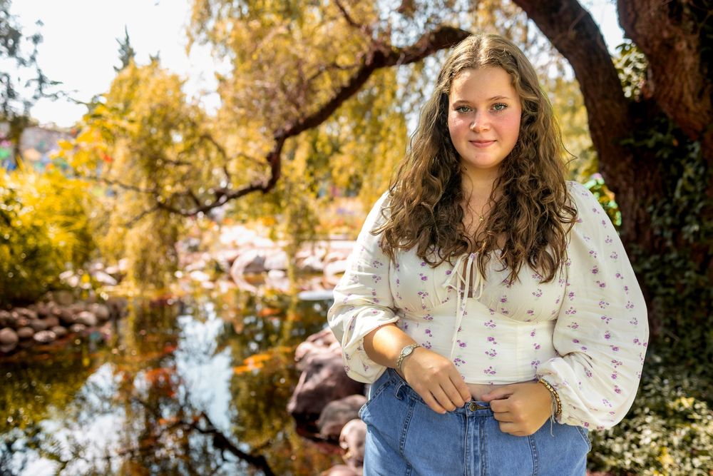 Person in peasant blouse and jeans sits near autumn pond with golden trees reflecting in water.