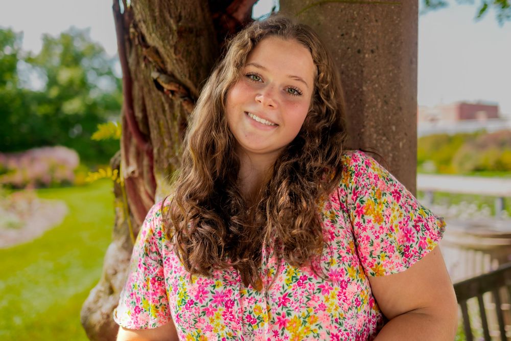 A person in a pink floral dress poses outdoors on a sunny day in front of a lush green background.