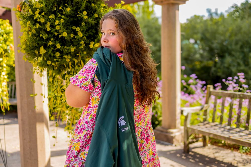 A long-haired figure in a floral dress and green scarf enjoys a sunlit garden setting with blooming flowers and pillars.