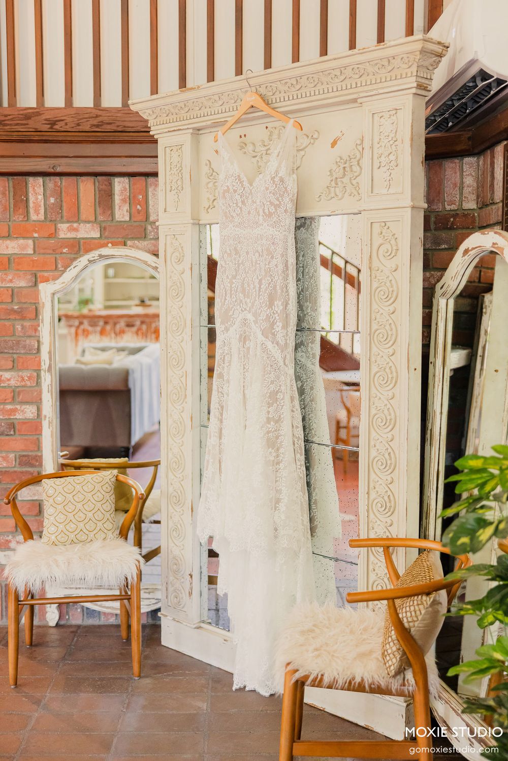Elegant white lace wedding dresses hanging on display in a rustic brick venue with wooden chairs.