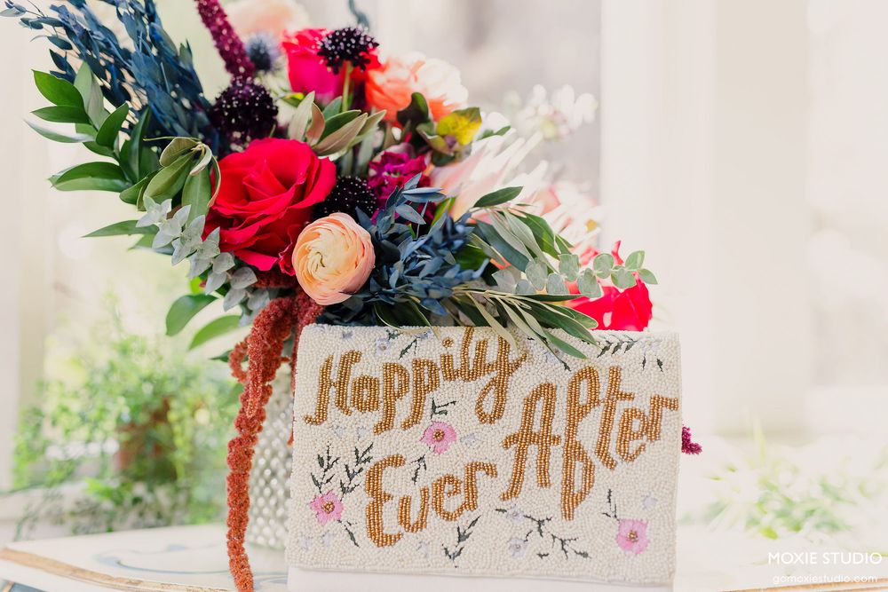 A rustic floral arrangement with red roses and blue accents sits atop a box decorated with 'Happily Ever After' text.