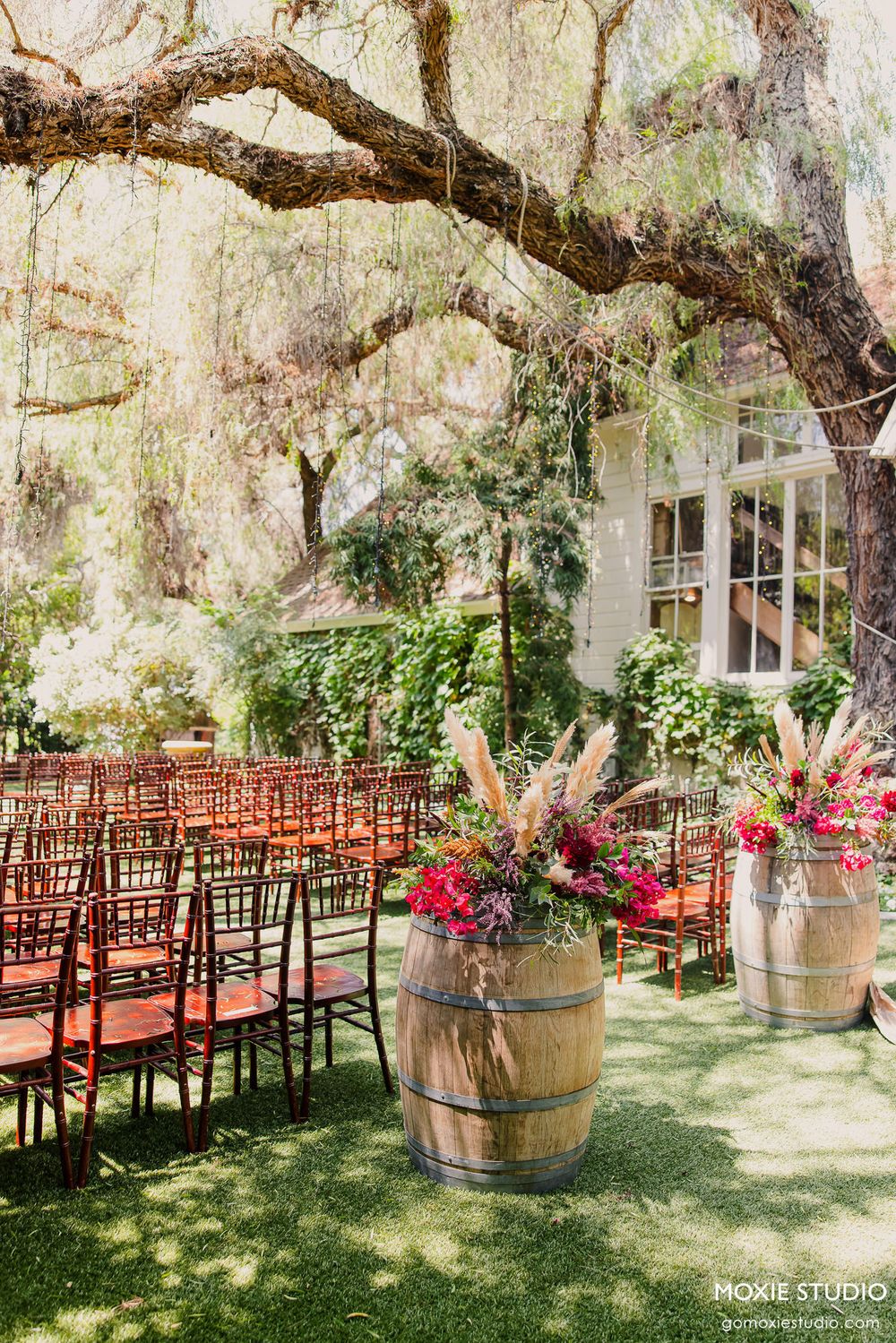 Rustic outdoor wedding ceremony setup with wooden chairs and wine barrel decorations under Spanish moss trees.