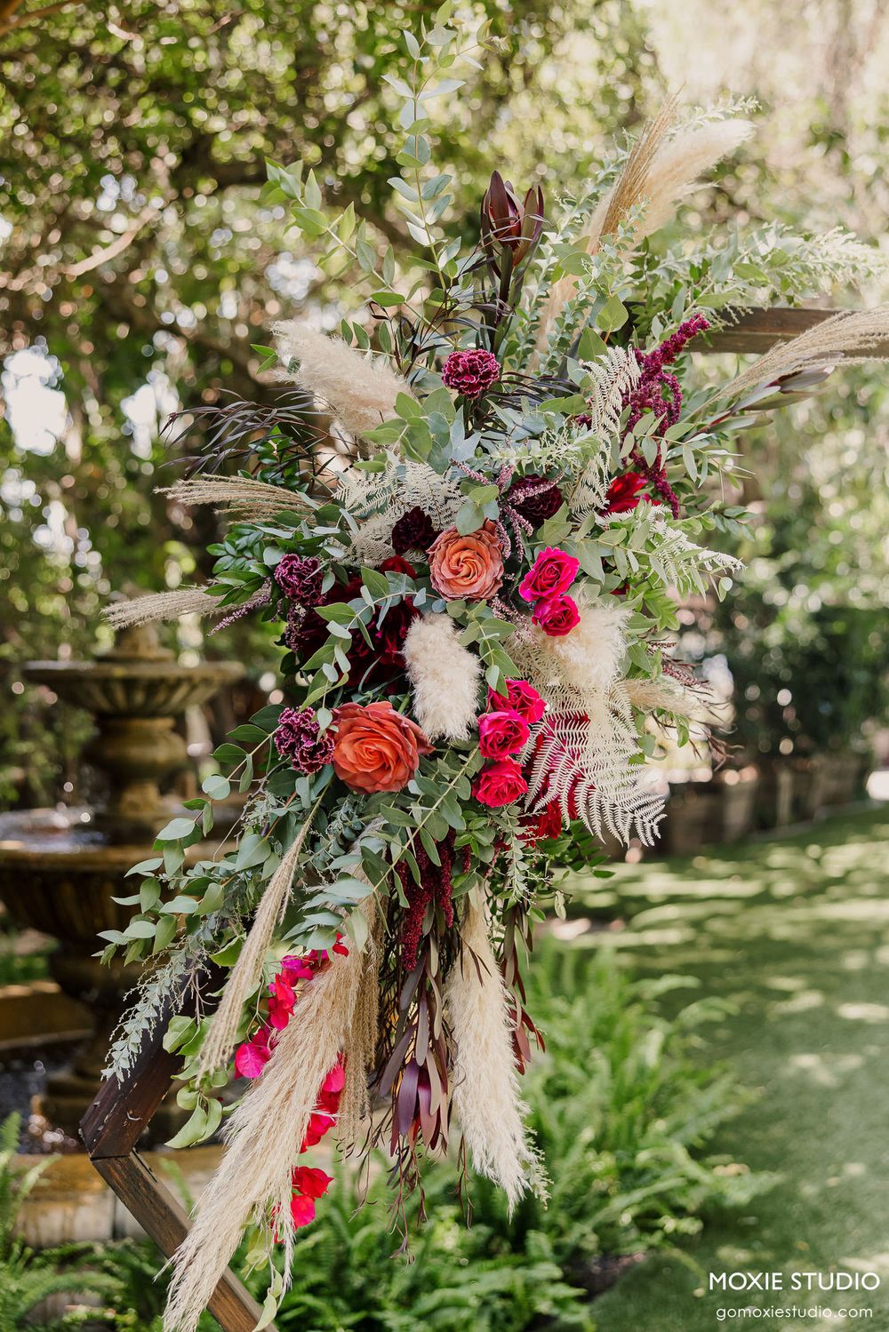 Lush floral arrangement with burgundy and pink blooms mixed with pampas grass and greenery in natural outdoor setting.