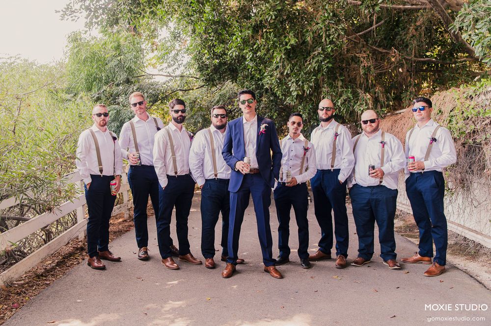 Wedding party members in blue suits and pink dresses line up outdoors for formal photos.