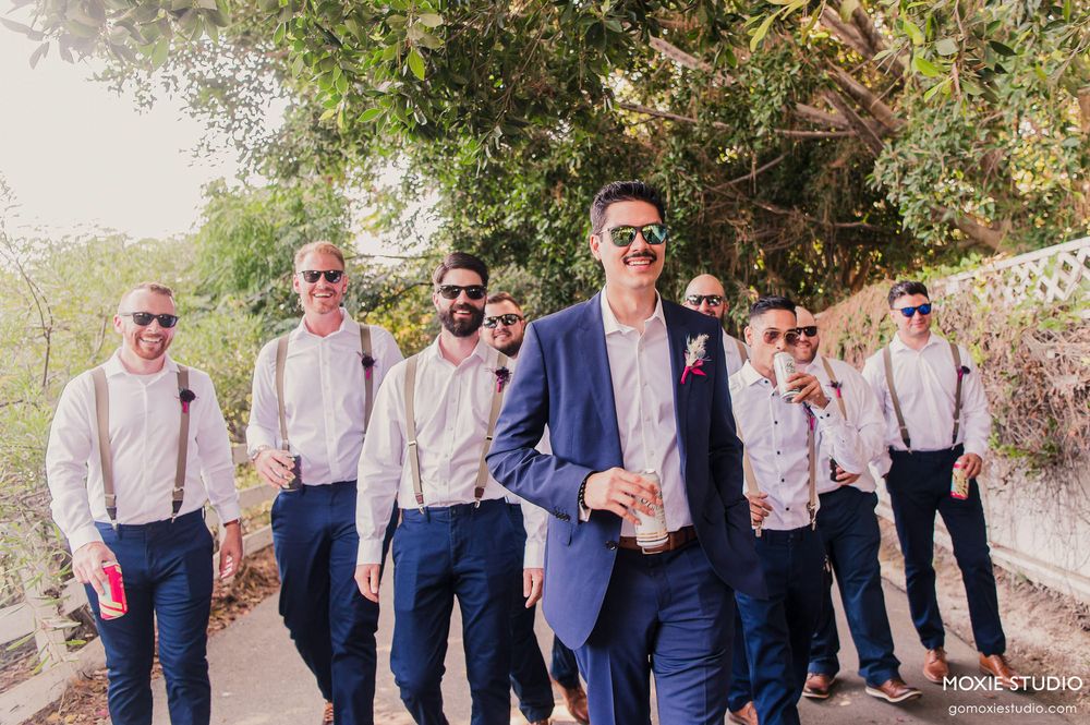 Groomsmen in navy suits and suspenders pose together outdoors on a sunny day.
