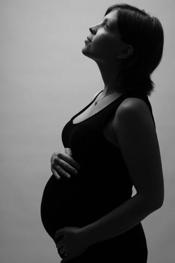 Black and white silhouette portrait of a pregnant woman in a black dress looking upward while holding her belly.