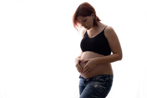 Pregnant woman in a black sports bra and jeans gently cradles her bare baby bump against a white background.