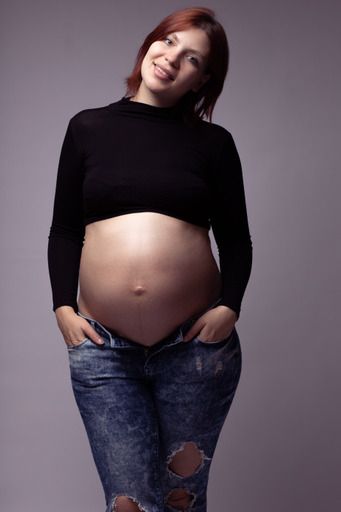 Smiling pregnant woman with red hair wearing a black crop top and ripped jeans showing her bare baby bump.