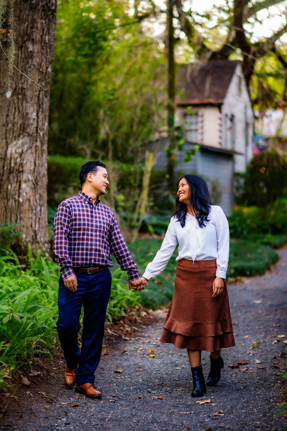 A couple holds hands while walking down a path near a white house in a lush garden setting.