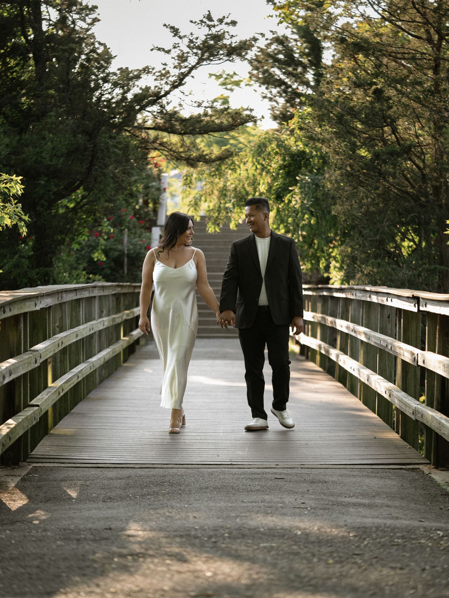 A pair in formal attire stroll together on a rustic wooden boardwalk through nature.