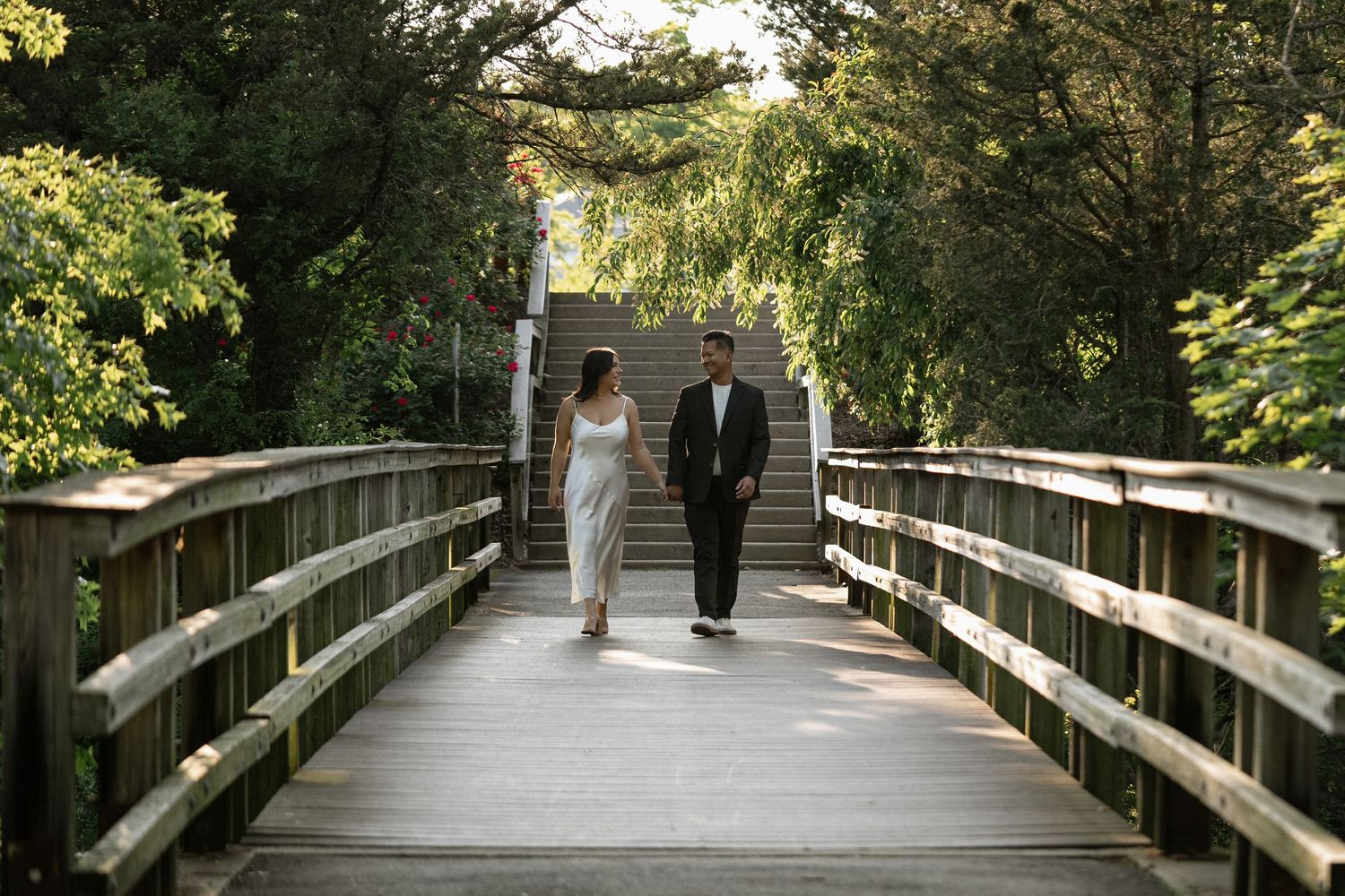 A couple walks along a wooden bridge pathway surrounded by lush green trees.