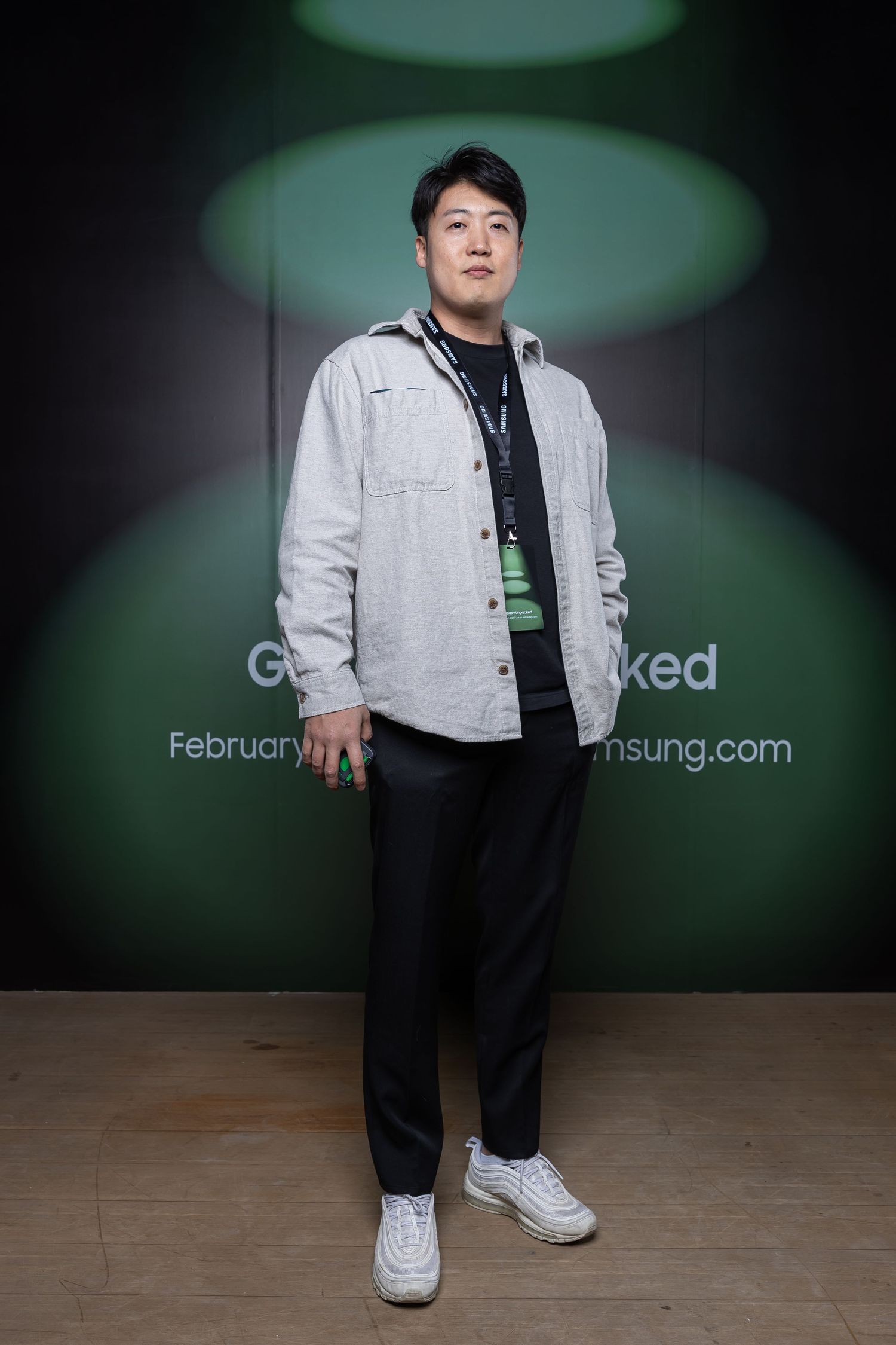 Person in light gray jacket and black pants standing against green promotional backdrop.
