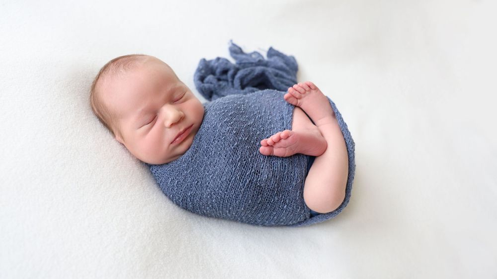 Newborn boy in a dark blue wrap posed sleeping. In-home newborn session with bump birth and beyond. Light hair soft skin.