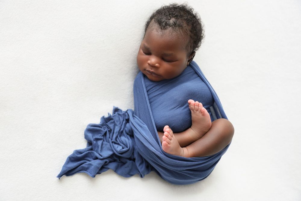 Peacefully sleeping newborn with tight curly dark hair and flowless soft skin, captured in natural light. Bue wrap featuring his tiny toes.