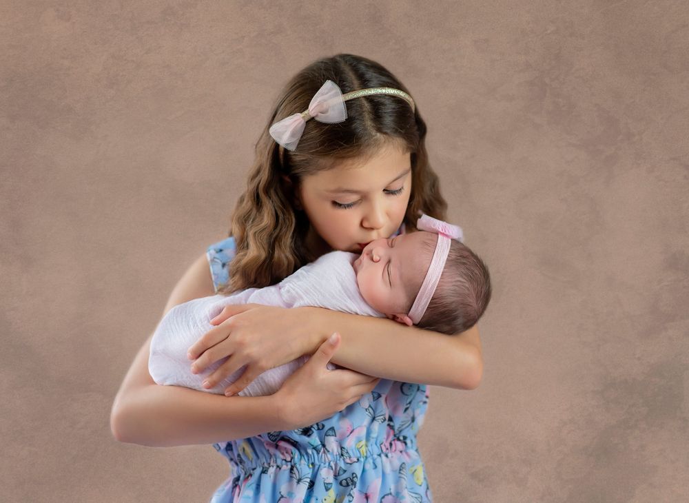 Sibling photo proud big sister holding her newborn Sister as she arrived home from the hospital. Kissing her cheek. Bump Birth and Beyond