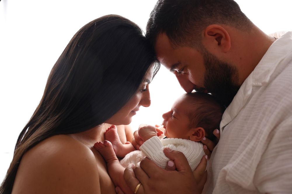 Newborn photoshoot, Penrith and surrounding suburbs. newborn boy in white set up with his parents for a backlit shot. Dad kissing newborn