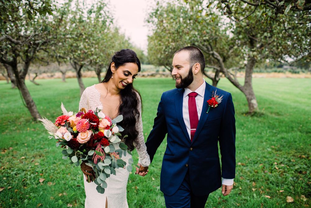Wedding couple walking through an orchard with bride holding red and pink bouquet.