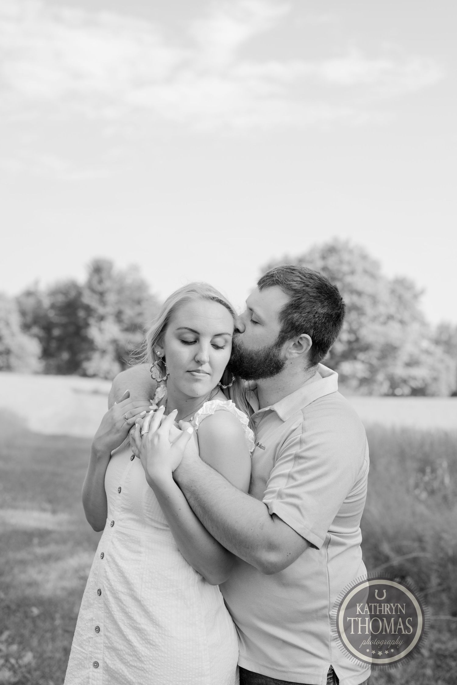 A tender black and white portrait of a couple embracing in a pastoral setting.