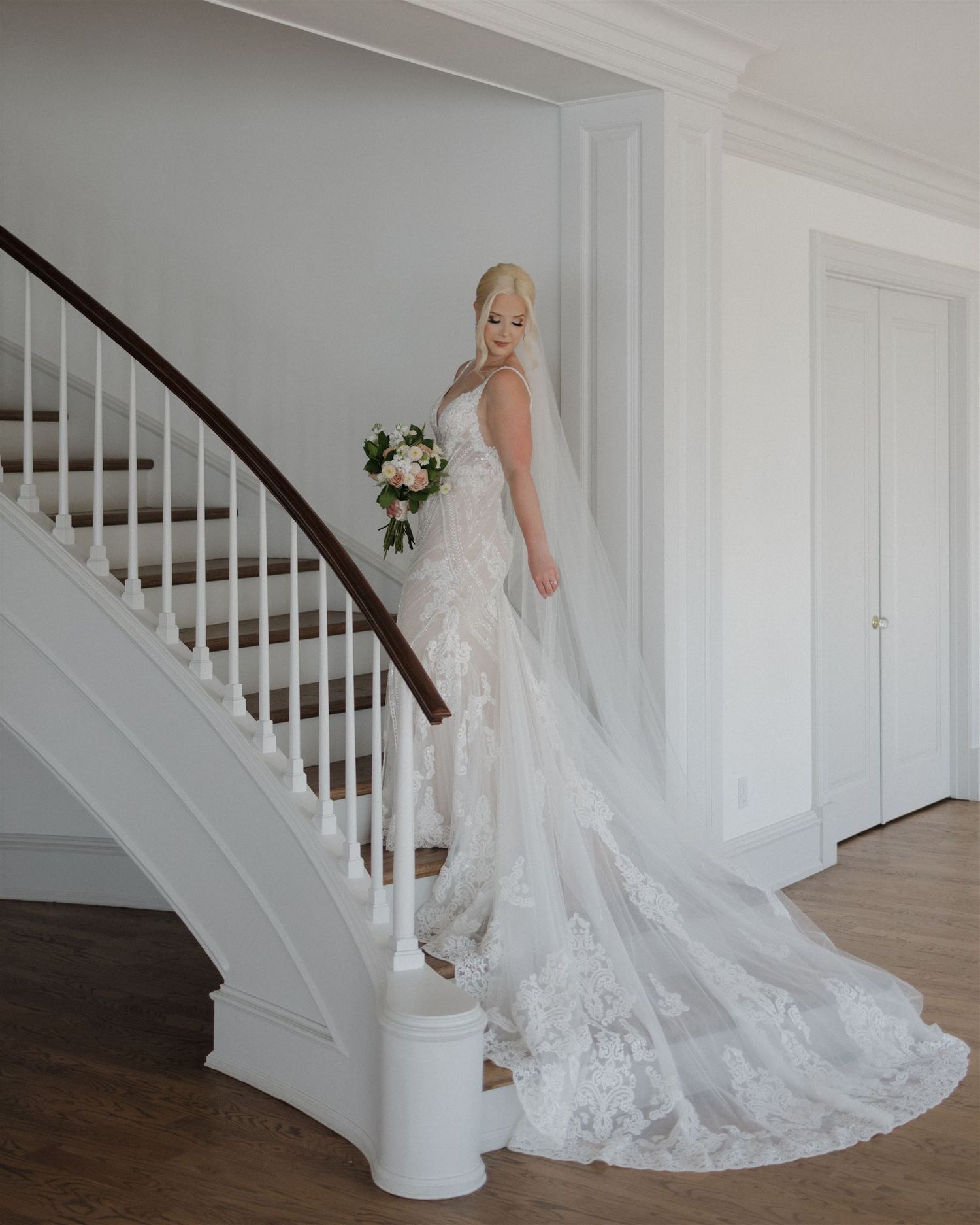 Full-length bridal portrait showcasing fitted lace gown with cathedral train on curved staircase.