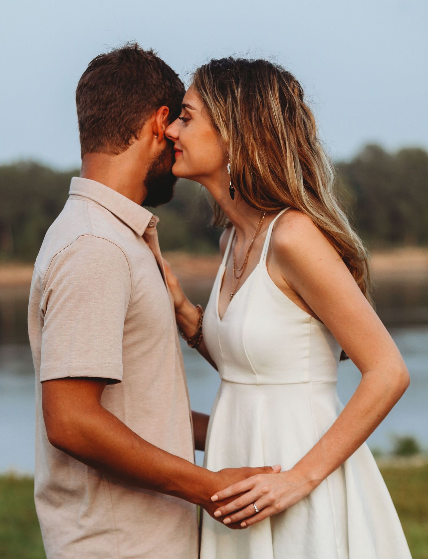 Romantic couple in white dress and beige suit sharing intimate moments by lake.